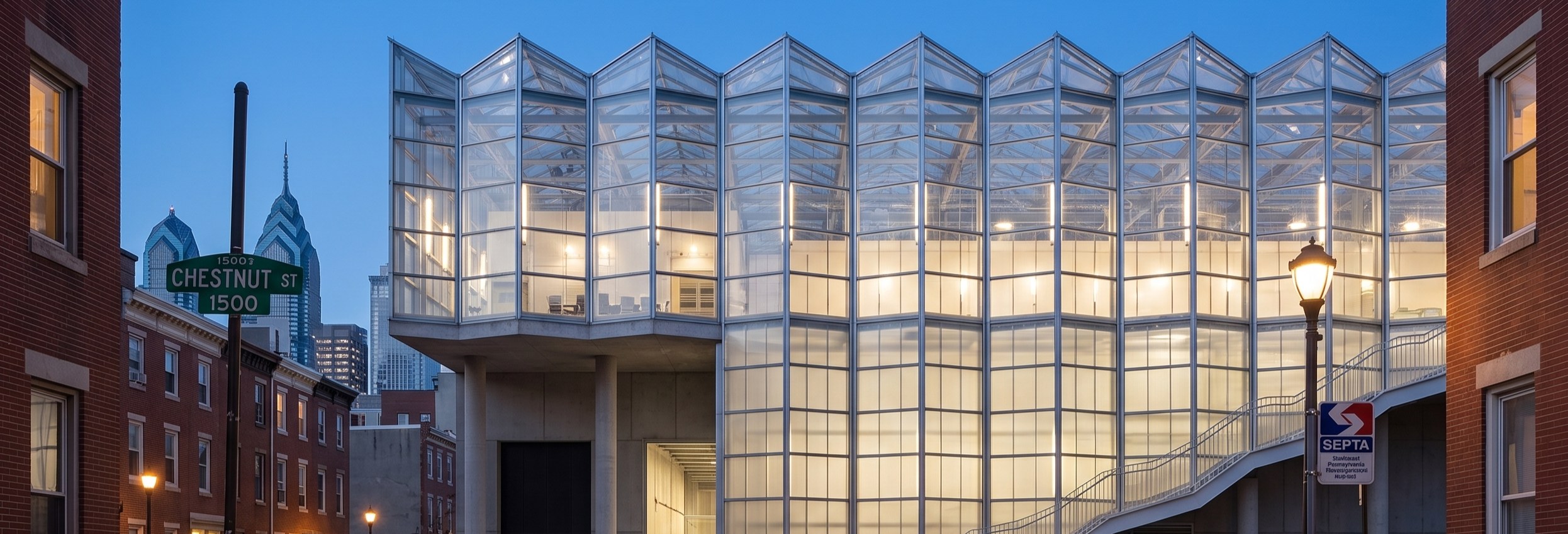 a glazed sawtooth roofed building lit from within at dusk, cantilevered over a city street between brick row houses.